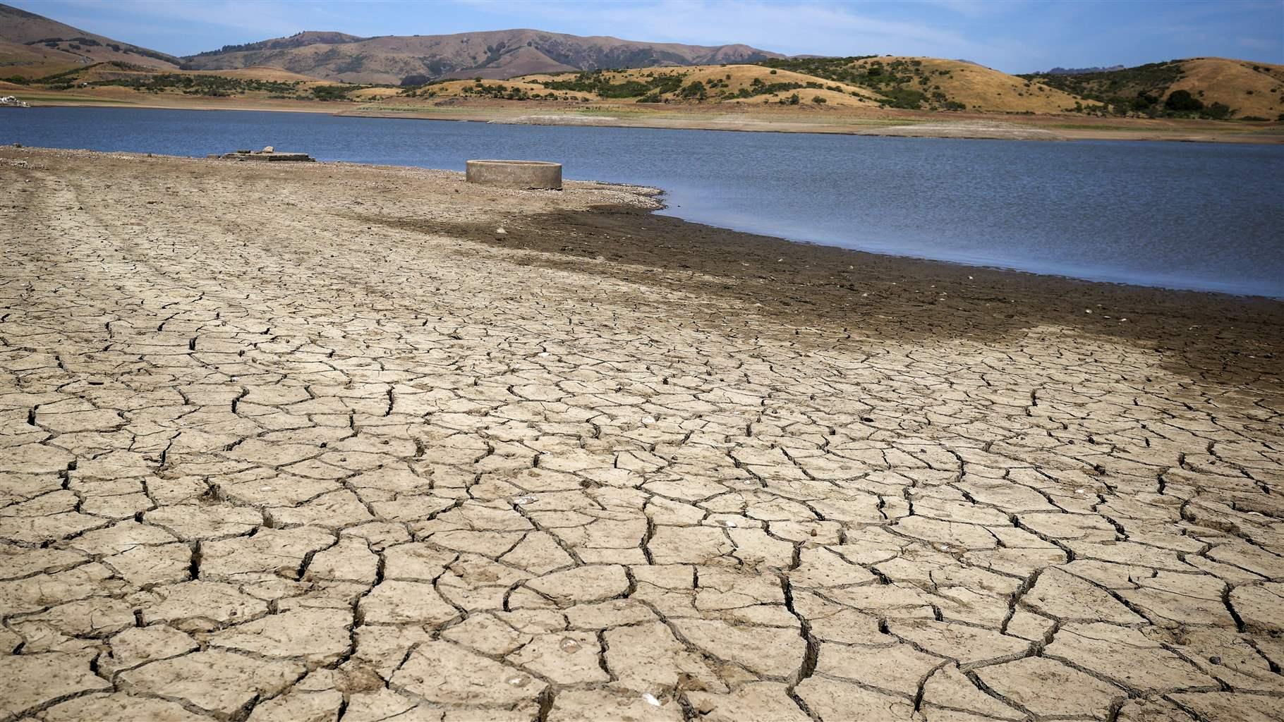 One part of the lake is dry and very cracked, and there is mud visible between the dry land and the water of the receding lake. Low desert hills in the background, very light clouds in the blue sky above.  