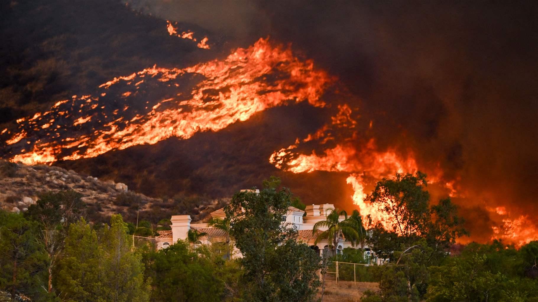 In the night picture, a wildfire burns across a low mountain and a steep slope behind the settlement.