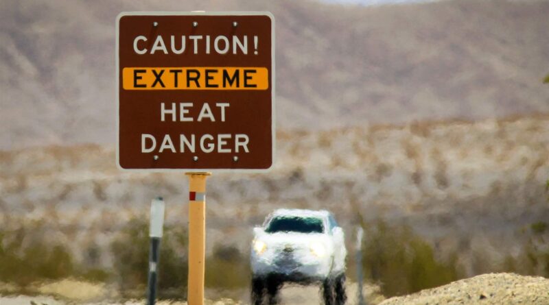 A road sign in the desert warns, in all capital letters, “Caution! Extreme heat risk.” The white car in the background has beige desert hills below. The whole surface is blurred by the camera settings and the fact that the heat is coming from the sand-and-asphalt area.